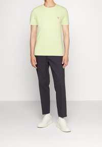 Svetle zelené tričko s krátkým rukávem a malým červeným logem, kombinované s tmavými kalhotami a bílými teniskami, stojící na světlém pozadí.