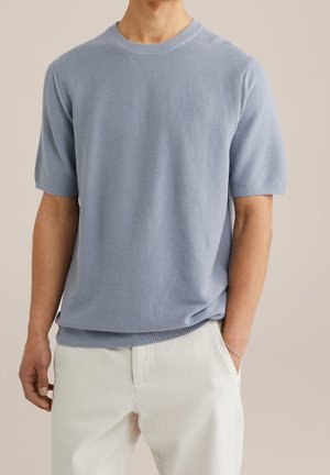 Man wearing light blue short-sleeve knit sweater and off-white pants, standing with one hand in pocket against neutral background.