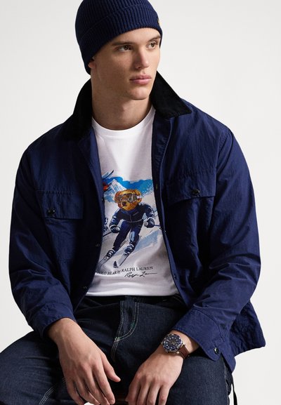 Mornarska jakna s crnim ovratnikom, bijela grafička majica s dizajnom medvjeda na skijanju, traperice i smeđi sat sa plavim brojčanikom.