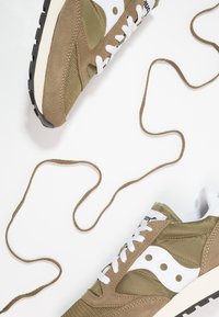 Hnědé semišové a textilní tenisky s bílými akcenty, kulaté tkaničky a texturovaná gumová podrá�žka, vystavené na bílém pozadí s volně uspořádanými tkaničkami.