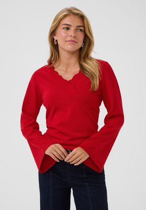 Femme aux longs cheveux blonds portant un pull rouge à col en V avec des bords festonnés et des manches cloche, associé à un jean foncé, sur un fond uni.