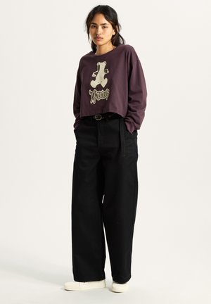 Cropped purple long-sleeve shirt with a bear graphic and logo, paired with high-waisted black wide-leg pants and white sneakers.
