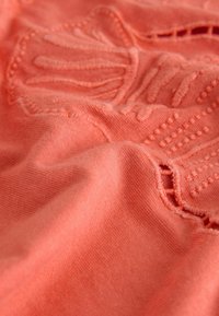 Korallivärvi kangas tekstuurse lillemustriga tikandiga. Pehme, sile tekstuur, kerge materjal. Nõrgalt läikiv pind toob esile kujunduse detailid.