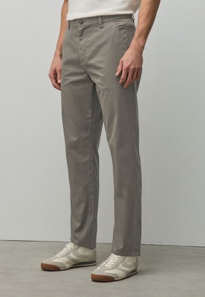 Mann trägt graue, gerade geschnittene Hose mit Seitentaschen und weiße Sneakers und steht auf einem glatten Boden vor einer schlichten Wand.