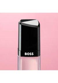 Blank sort kasket med hvid "BOSS" tekst på en klar parfume flaske mod en blød lyserød baggrund.