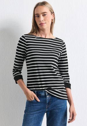 Femme aux longs cheveux blonds portant un t-shirt rayé noir et blanc à manches longues et un jean bleu, debout avec une main dans la poche.