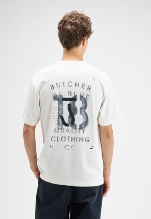 Homme aux cheveux bouclés portant un t-shirt blanc avec un texte noir "Butcher of Blue Quality Clothing Co", vu de dos.