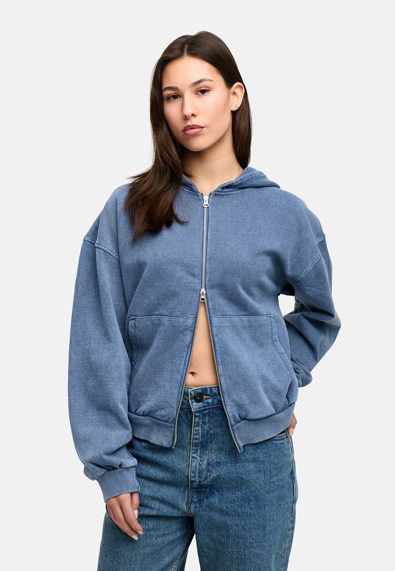 Blue zip-up hoodie made of soft fabric with a hood, dropped shoulders, and ribbed cuffs. Paired with high-waisted blue denim jeans.