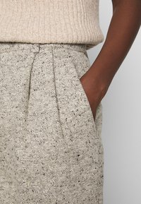 Pantaloni in tessuto beige chiaro testurizzato con chiazzette nere. Presentano una piega in vita e una tasca laterale, che si abbinano a un top cremoso a coste.