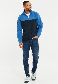 Fleece-Pullover in Hellblau und Marineblau, mit einem halben Reißverschluss und gesticktem Logo. Kombiniert mit dunklen Jeans und sportlichen Schuhen. Glatte Textur.