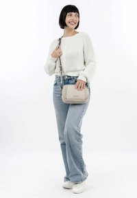 Donna con capelli neri corti, che indossa un maglione bianco, jeans azzurri chiari e scarpe da ginnastica bianche, mentre tiene una borsa a tracolla beige, sorridente, in piedi su sfondo bianco.
