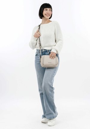 Donna con capelli neri corti, che indossa un maglione bianco, jeans azzurri chiari e scarpe da ginnastica bianche, mentre tiene una borsa a tracolla beige, sorridente, in piedi su sfondo bianco.
