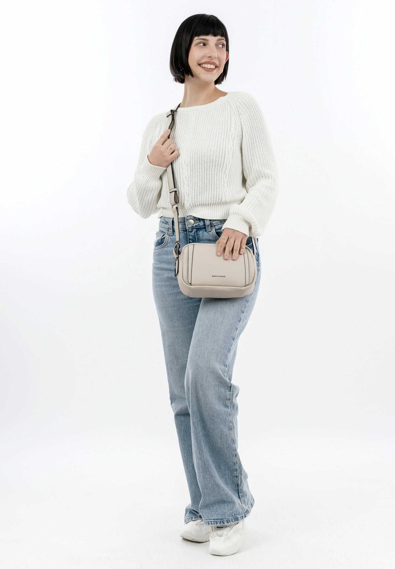 Donna con capelli neri corti, che indossa un maglione bianco, jeans azzurri chiari e scarpe da ginnastica bianche, mentre tiene una borsa a tracolla beige, sorridente, in piedi su sfondo bianco.