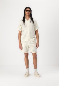 Camicetta a righe beige a maniche corte e pantaloni corti abbinati, realizzati in un tessuto morbido. Abbinati a sneakers di colore chiaro e occhiali da sole.