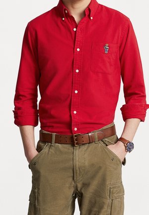 Mężczyzna w czerwonej koszuli zapinanej na guziki z emblematem niedźwiedzia na kieszeni, podwiniętymi rękawami, brązowym paskiem, spodniach typu cargo oraz zegarku na rękę z niebieską tarczą.