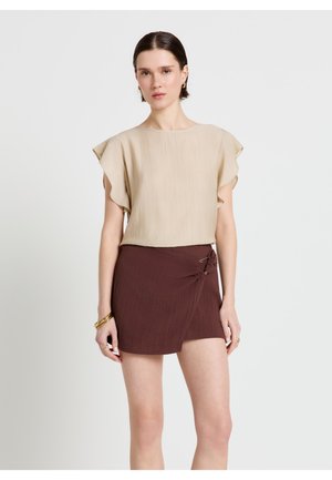 Frau mit kurzen dunklen Haaren, die ein beigefarbenes Oberteil mit flatternden Ärmeln und einen dunkelbraunen Wickelrock trägt, steht vor einem schlichten hellen Hintergrund.