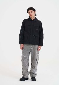 Młoda osoba z włosami do ramion, ubrana w czarną kurtkę z kieszeniami oraz szerokie szare dżinsy z haftowanymi wzorami i czarne buty.