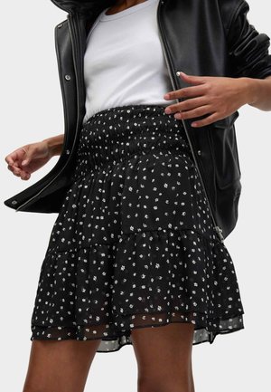 Frau trägt eine schwarze Lederjacke über einem weißen Oberteil und einem schwarzen Rock mit kleinen weißen Blumenmustern und hebt leicht die Kanten der Jacke an.