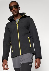 Svart softshell-jakke med gule glidelåser, glidelåslommer og høy krage. Tøyet ser glatt ut og har en lett struktur.