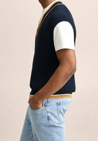 Mann trägt eine navyblaue Strickweste mit gelben und weißen Akzenten über einem weißen kurzärmligen Hemd, hellblaue Jeans, und steht mit einer Hand in der Tasche.