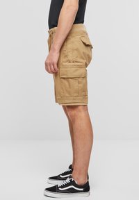 Beige cargoshorts med sidelommer og baklommer. Laget av robust stoff, med en avslappet passform og elastisk midjebånd. Brukes med svarte sneakers.