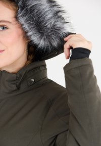 Mörkgrön jacka med en foder som är klädd i päls, med hög krage och tryckknappar. Tyget har en slät textur och pälsen är grå.
