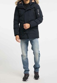 Marinblå puffjacka med pälsfodrad huva, utrustad med tryckknappar, sidofickor och en armficka, kombinerad med ljusblå jeans och sneakers.