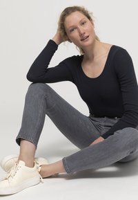 Långärmad marinblå topp med rundad halsringning, grå skinny jeans och ljusfärgade sneakers med knutna snören, sittande framför en enkel bakgrund.