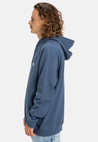 Sudadera con capucha de tela azul oscuro, con un parche de logo en el frente, mangas largas y un corte relajado. Textura suave y diseño clásico.