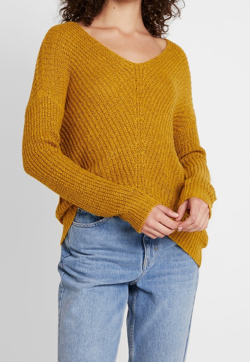 Femme portant un pull en maille texturée jaune moutarde avec un col en V et un jean taille haute bleu clair, mains jointes devant.