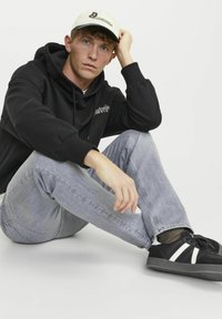 Sweat à capuche noir, jeans gris clair et baskets noires avec des rayures blanches. Casquette de couleur claire avec des détails graphiques. Fond neutre.