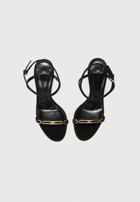 Black suede high-heeled sandals with thin ankle straps, featuring a gold chain accent across the toe and branded insole.