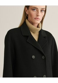 Sort uldblandingsfrakke med dobbeltbrystet design, notched lapels og to knapper; parret med en lysbeige ribbet turtleneck sweater.
