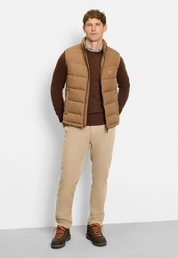 Braune Steppweste über einem braunen Pullover, kombiniert mit beigen Hosen. Braune Stiefel mit orangefarbenen Schnürsenkeln. Stehend vor einem weißen Hintergrund.