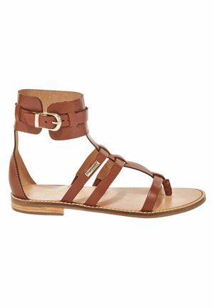 Sandale en cuir marron avec bride de cheville, semelle plate, design ouvert aux orteils et plusieurs lanières sur le pied. Présente un détail de boucle en or.