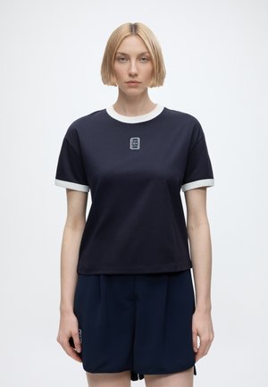 Femme blonde aux cheveux courts, debout face à l’avant, portant un t-shirt bleu marine et un short avec des bordures blanches aux manches et au col.