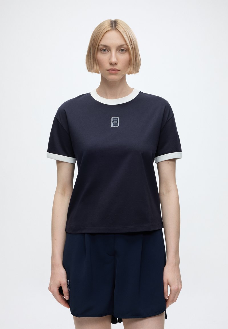 Femme blonde aux cheveux courts, debout face à l’avant, portant un t-shirt bleu marine et un short avec des bordures blanches aux manches et au col.