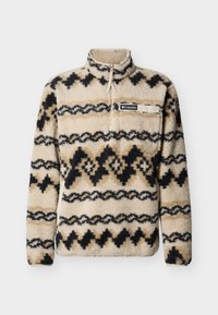 Μπεζ φλις μπλούζα με ασπρόμαυρα και καφέ γεωμετρικά σχέδια, ημι-φερμουάρ στον λαιμό και τσέπη στο στήθος με λογότυπο. Απαλό, υφασμάτινο υλικό με υφή.