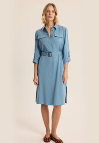 Jasnoniebieska sukienka koszulowa z długimi rękawami, zapinana na guziki z przodu, posiadająca dwie kieszenie na piersi oraz pasek w talii. Charakteryzuje się prostym krojem i rozcięciami po bokach.
