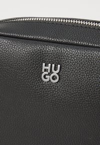 Black textured leather bag with a silver "HUGO" logo. Features a metal zipper along the edge and a smooth finish.