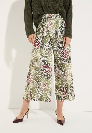 Woman wearing wide-leg pants with green, white, and red abstract leaf pattern and a green knit sweater standing on white floor.