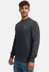 Dunkelgrauer Sweatshirt mit rundem Halsausschnitt und gerippten Bündchen. Verfügt über ein kleines Logo auf der Brust. Kombiniert mit blauen Jeans.