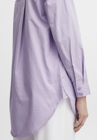 Lavender long-sleeve shirt made of lightweight fabric, featuring a curved hem, buttoned cuffs, and a relaxed fit. Smooth texture.