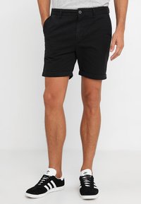Schwarze Baumwollshorts mit umgeschlagenen Säumen, mit einem Knopfverschluss und seitlichen Taschen. Getragen mit schwarz-weißen Sneakers.
