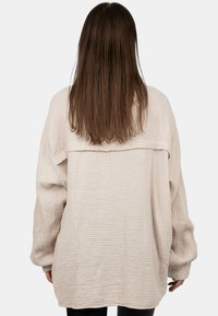 Lichtbeige, textuurrijke oversized shirt met lange mouwen en een verlaagde schoudernaad. Heeft een horizontale naad op de rug.