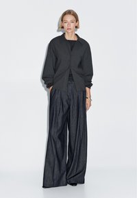 Black oversized shirt with a collar, paired with wide-legged dark denim trousers. Model stands facing forward against a plain background.