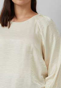 Donna che indossa una blusa a maniche lunghe di colore crema, con texture, scollatura rotonda e maniche leggermente a sbuffo.