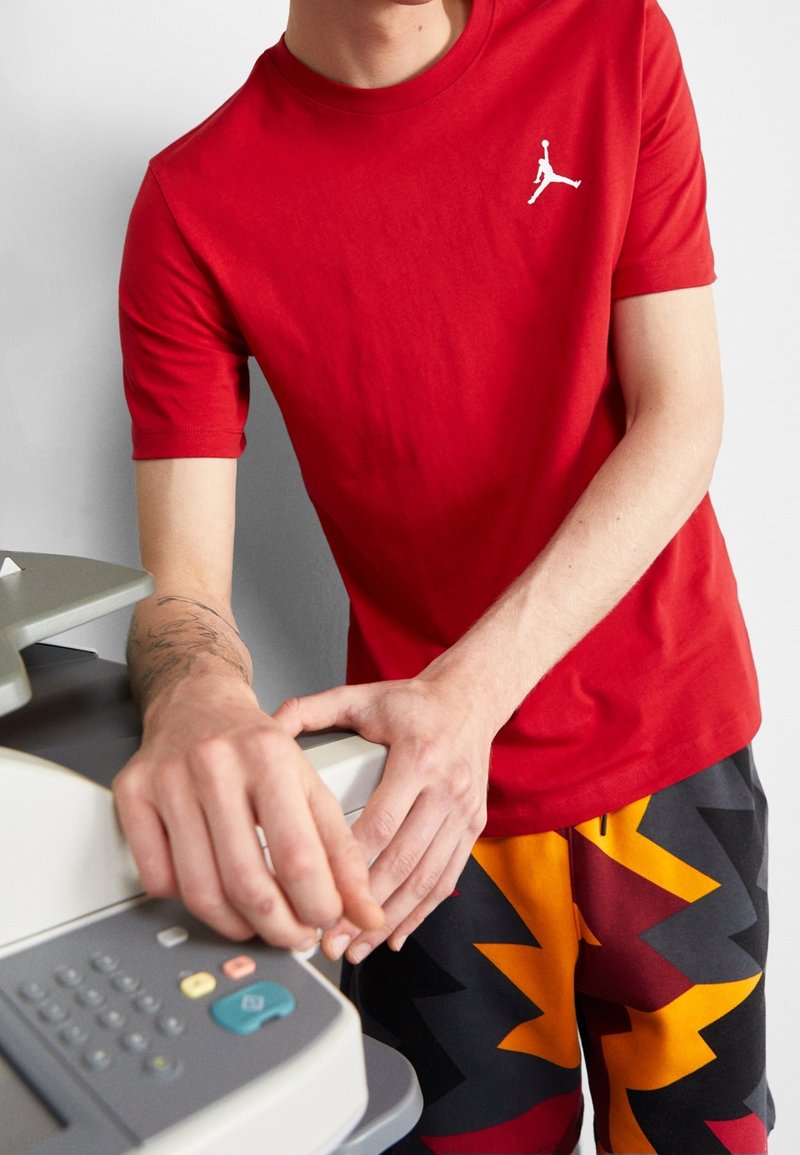 Persona vestida con una camiseta roja de Jordan y pantalones cortos de patrón zigzagueante colorido, operando una fotocopiadora o impresora gris con una mano sobre la máquina.