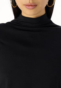 Svart høyhalset topp laget av glatt, elastisk stoff. Den har en tettsittende design uten synlige sømmer, og er komplementert med store gullhårøredobber.