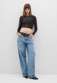 Schwarzes Pailletten-Crop-Top, hoch taillierte hellblaue Jeans mit weitem Bein, schwarzer Gürtel mit rundem Schnallenverschluss, spitze schwarze Absätze, stehende Pose vor einer weißen Wand.